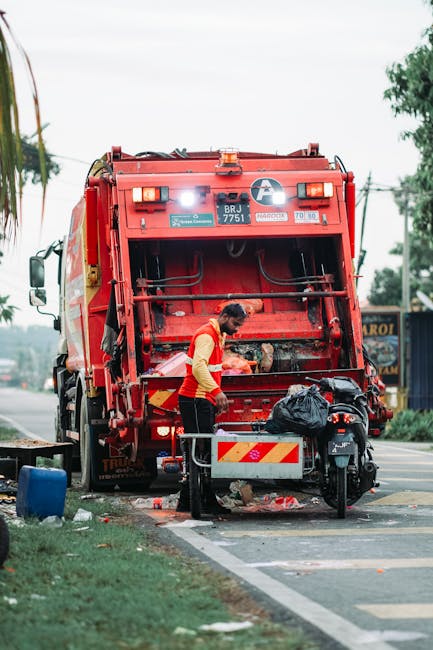 A waste collection worker in a high-visibility orange and yellow jacket is operating a large red rubbish collection truck on a city street. The truck's rear compactor section is open, revealing the in