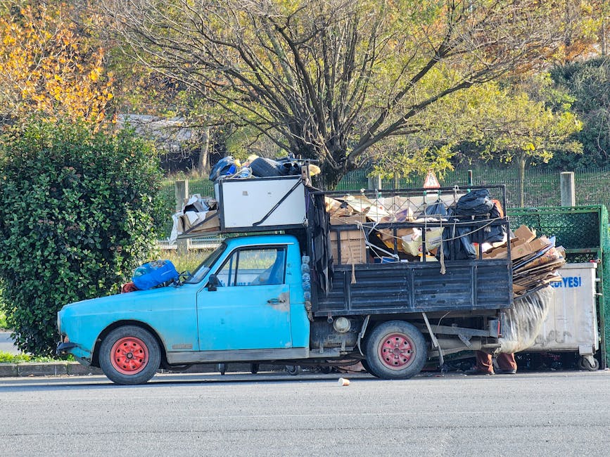 A waste collection worker in a high-visibility orange and yellow jacket is operating a large red rubbish collection truck on a city street. The truck's rear compactor section is open, revealing the in