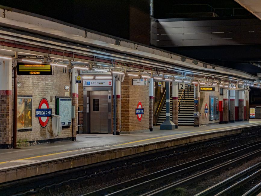 A nighttime view of a London Underground train station platform showing brick walls with worn, beige-colored tiles and signage indicating Harrow & Wealdstone station. The platform is illuminated by ov